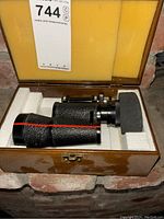Single lens binocular placed inside a wooden box with foam padding, showing the full binocular and case interior.