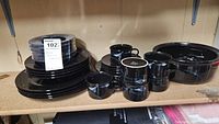 Complete view of black Mikasa dishware including plates, bowls, and coffee cups arranged on a shelf.