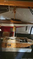 Photo showing a model boat with orange patterned hull marked 1088 with motor, and wooden boat hull on shelf above