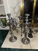 Pair of silver-tone five-arm candelabras with detailed bases and arms, placed on marble surface near a fireplace, also visible nearby metal candle stands.