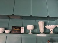 Three small white milk glass footed items including a round bowl, hexagonal vase, and scalloped candy dish on a teal shelf