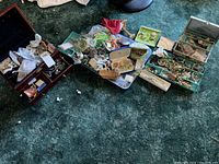 Overview of multiple jewelry boxes, trays, and loose costume jewelry pieces including earrings, rings, bracelets, and necklaces scattered on carpet.