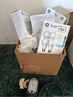 Box containing multiple packages of GE Basic LED 750 lumens lightbulbs and some packing materials on dark floor.
