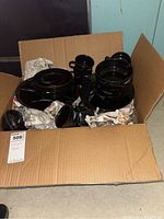 Overview of the open cardboard box containing black glass plates, cups, and other dishware wrapped partially in newspaper.