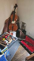 Full view of cello leaning against wall with soft case behind, cello bow across strings, assorted percussion on floor