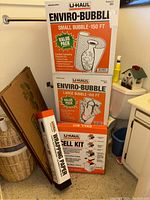Photo showing stacked boxes of U-Haul Enviro-Bubble Small and Large Bubble Wrap and a Cell Kit in a corner with other household items.