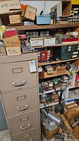 Full view of the 4 drawer beige metal file cabinet alongside shelving with various small hardware items in boxes and drawers.