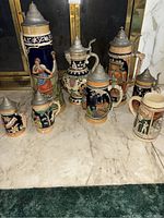 Photo shows seven assorted ceramic beer steins with pewter lids displayed on marble surface in front of a fireplace. Various hand-painted traditional German motifs and colors are visible.