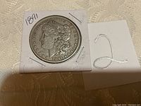 Front obverse side of 1891 Morgan silver dollar coin showing Lady Liberty profile, year 1891, and inscriptions 'E PLURIBUS UNUM' with 13 stars around the edge in a white cardboard holder.