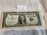 Front of 1935 E silver certificate one dollar bill in a clear protective sleeve on a textured surface, with a paper tag marked 55.