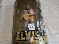 Elvis Presley doll in packaging holding guitar standing beside microphone stand, displayed on white patterned surface.