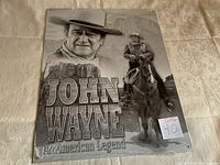 Front image of John Wayne replica tin sign showing grayscale images of John Wayne and text 'John Wayne An American Legend', sealed in plastic.
