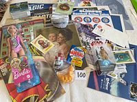 Photo of assorted items including Barbie doll, trading card packs, collectible coins, vintage poster, and Star Wars figure