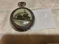 Front view of silver-tone pocket watch showing black and white detailed image of a steam train on the cover.