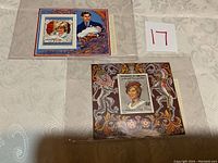 Two commemorative stamp sheets in protective sleeves placed on a table. One shows Prince Charles with baby Prince William and Princess Diana's stamp, the other shows a large stamp of Princess Diana.