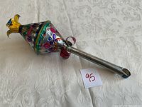 Vintage decorative metal object placed on white patterned fabric, showing colorful hand-painted floral designs and a silver long handle with a rounded end.