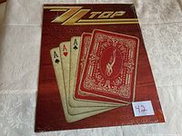ZZTop tin sign replica sealed with a tear in the plastic wrapping on the bottom corner, front view showing playing card design with aces and ZZTop logo on wood grain background.