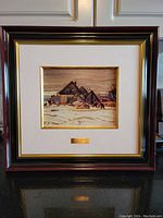 Frontal image of the framed lithograph showing the dark brown wooden frame with gold trim and cream matting around the winter village scene.