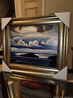 Framed lithograph artwork showing landscape with cloud formations and lake in blues and dark colors. Frame has protective corners.
