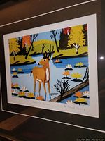 Framed print showing colorful folk art deer in stream with flowers and autumn trees, title and artist name signed on print.