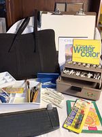 Overview of various art supplies including black padded carrying case, white paint palette, pads of watercolor paper, and carrying box with paint tubes.