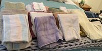 Photo showing multiple folded bath towels in cream, beige, white, lavender, brown, and pastel striped patterns arranged on a bed.