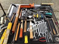 Overview of hand tools displayed on black surface including hammers, wrenches, screwdrivers, measuring tape, and other tools laid out neatly.