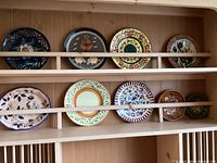 Full view of 8 handpainted ceramic plates arranged on a wooden shelf with front protective bars, showing a variety of colorful designs and shapes.