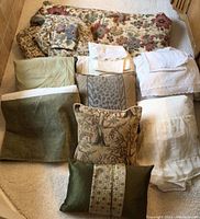 Photo showing assortment of bed linens including floral blanket, multiple folded sheet sets in white, beige, and cream tones, and assorted pillows in neutral and green colors.