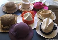 Overview of eight ladies summer hats arranged on a dark surface showing variety of styles, materials, and colors.