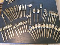 Full layout of all flatware pieces displayed on black fabric showing knives, forks, spoons and serving pieces with decorated handles.