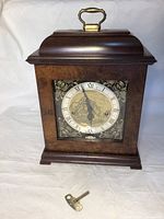 Front view of entire clock showing wood case, brass handle, and glass door with clock face visible.