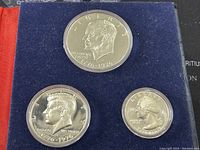Obverse side images of the silver dollar, half dollar, and quarter coins showing the profiles of Liberty, Kennedy, and Washington respectively with the dates 1776-1976.