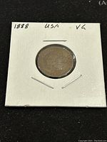 Obverse side of the 1888 Indian Head penny, shows Lady Liberty with Native American headdress, held in white cardboard holder.