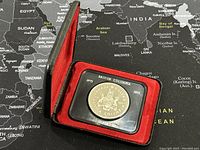 Coin displayed inside protective plastic case and red-lined display box on a world map background.