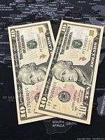 Two US $10 banknotes displayed showing the front side with Alexander Hamilton's portrait and visible design elements.