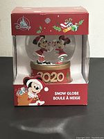 Front view of the 2020 Disney Mickey and Minnie Mouse Christmas snow globe in original red holiday packaging, showing the snow globe inside a clear plastic window box.