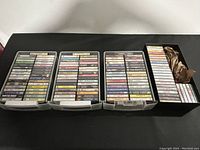 Four trays and a box filled with over 130 cassette tapes organized in rows, displaying cassette cases with visible printed labels.