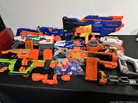 Full view of the assortment of Nerf blasters and accessories laid out on a black surface. Multiple blasters of various sizes and colors including orange, blue, white, green, and black. Several detachable magazines and scoped attachments visible alongside a bag of darts.