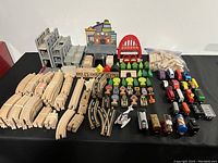 Overview image showing large assortment of wooden train tracks, train cars, buildings, trees, and road sign accessories organized on a black table surface.