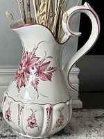 Close-up view of ceramic ewer showing red floral design, curved spout, and handle on a white base.
