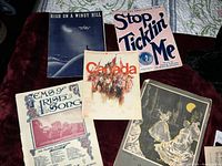 Five vintage sheet music covers spread on a dark red velvet surface, titles visible include 'High on a Windy Hill', 'Stop Ticklin' Me', 'Songs of Irish Song', 'Canada Centennial Song' and a black and white illustration cover.