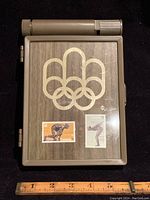 Brown case with wood grain and gold Olympic rings showing two stamps: $1 depicting runner sculpture, $2 depicting diver sculpture