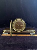 German wood mantel clock with open brass bezel showing large round dial with Roman numerals and wooden base with curved ends, measured against ruler for size.