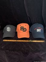 Three caps side by side on a black background with a measuring ruler in front; from left to right: navy blue San Diego cap, orange San Diego cap, navy blue slogan cap