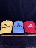 Three Cobra branded baseball caps in yellow, blue, and red, displayed side by side with a ruler measuring length in front of them for scale.