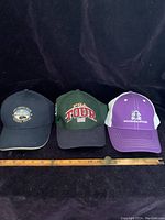 Three golf caps lined up side by side with a measuring stick in front showing each cap size; navy, green, and purple caps.