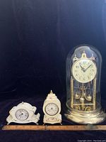 Three clocks arranged side by side with ruler placed in front showing size. Left: mini Westclox mantel clock with floral decoration. Center: taller Westclox desk clock also floral decorated. Right: Ergo anniversary clock under glass dome with gold base.