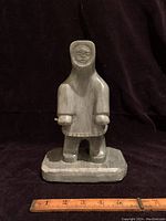Front view of a hand-carved soapstone seal hunter figure on a rectangular base with a ruler for scale showing approximately 9 inches high.
