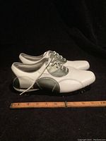 Side view of the pair of white and silver leather golf shoes with a ruler showing length scale.